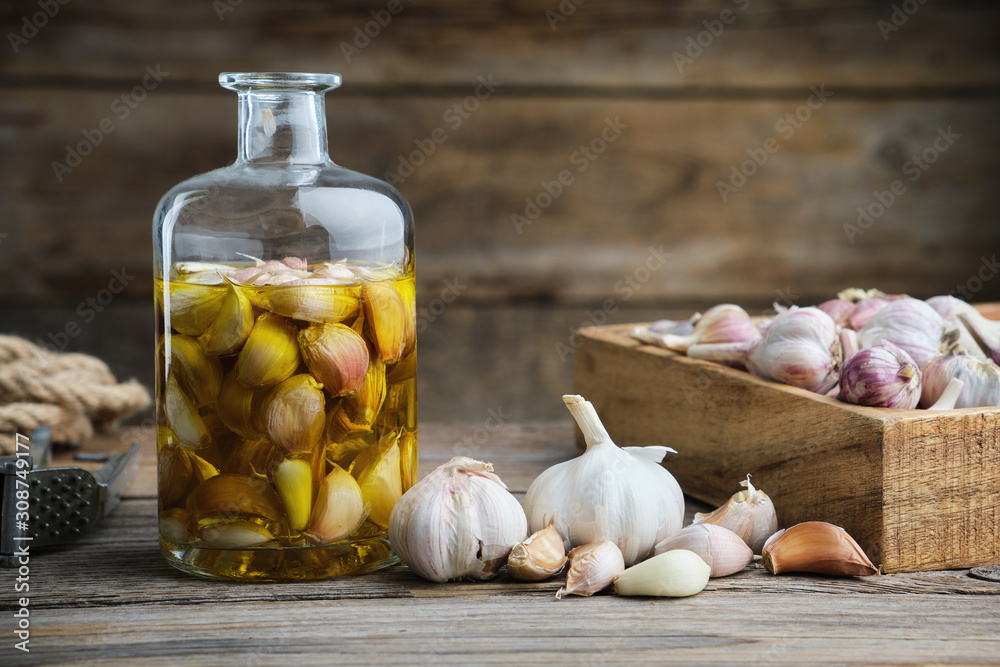 Garlic aromatic flavored oil or infusion bottle, wooden crate of garlic cloves and  garlic press on wooden kitchen table. - obrazy, fototapety, plakaty 