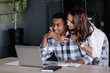© Rithor - Beautiful young woman in love hugs her positive successful african-american businessman husband working in the office at a table with a laptop and notebook. Loving mixed race couple concept