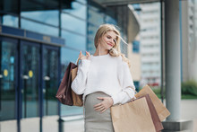 Young Attractive Pregnant Blonde Woman In A White Sweater Against The Backdrop Of The City Streets. Holding A Lot Of Shopping Bags.