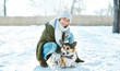 © vitaliymateha - Young happy woman in woolen hat and long warm scarf smiling to camera, stroking her pet and having fun in snowy winter park at frosty sunny day. Happy time together, cute dog Welsh Corgi Pembroke