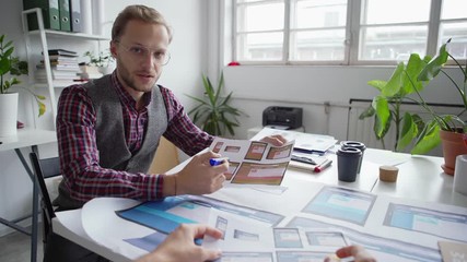 Wall Mural - POV of young entrepreneur in eyeglasses showing mobile application interface samples printed on paper to colleague sitting at desk in office