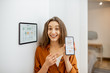 © rh2010 - Portrait of a young and happy woman near a touch screen panel for managing smart home. Holding smart phone with a simlpe and convinient mobile application for smart home
