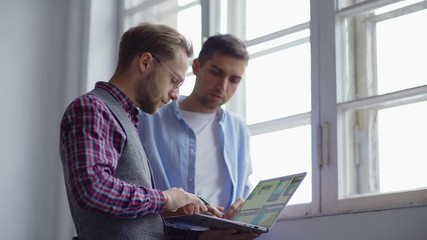 Wall Mural - Waist up of two young web designers standing by window at office and discussing app interface samples presented on laptop
