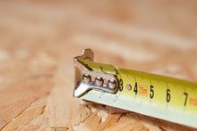 A Tip Of Yellow Metal Tape Measure Lying On Chip Board. Blurred Background.