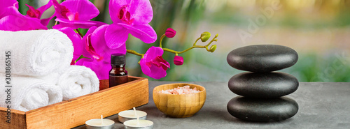 Fototapeta na szkło hot spa stones set for massage treatment, orchid flower, towels, candles and sea salt on green background with bamboo. Elegant and luxury spa. mock up, template. Health and beauty care concept