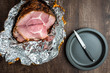 Spiral cut glazed and cooked ham in a foil wrapper on a wood table, plate and knife ready to cut ham