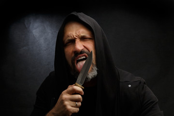 Portrait of a bald man with a beard in a black hood with a knife on a dirty gray background. Maniac.