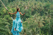 Tanned beautiful woman in a long turquoise dress with a train, riding on a swing. In the background, a rainforest and palm trees. Copy space