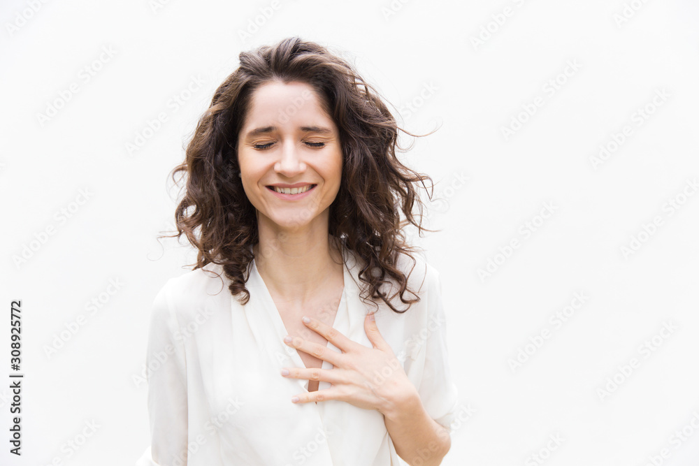 Happy grateful pretty woman with closed eyes applying hand to chest. Wavy haired young woman in casual shirt standing isolated over white background. Relief or gratitude concept - obrazy, fototapety, plakaty 