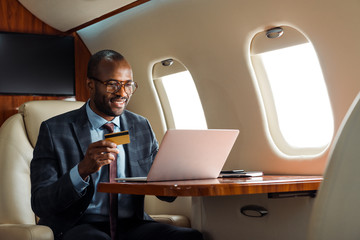 happy african american businessman in glasses holding credit card near laptop in private plane