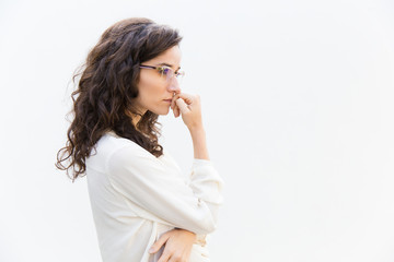 Side of pensive office assistant in glasses leaning face on hand and looking away at copy space. Wavy haired young woman in casual shirt standing isolated over white background. Advertising concept