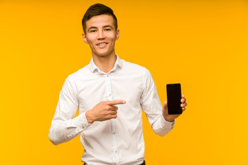 Excited winner Asian man pointing at mobile phone after received good news isolated on yellow background in studio.