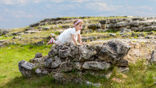Little Caucasian Girl In A Dress Scrambles On Old Outdoor Ruins Stones. Beautiful Elegant Child Walking In The Summer Beautiful Landscape Outdoor