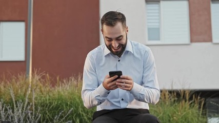 Wall Mural - Cheerful bearded businessman using smartphone while sitting on bench outdoors