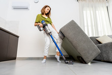Young Woman Cleaning Floor Under The Sofa With Cordless Vacuum Cleaner At Home. Concept Of Easy Cleaning With A Wireless Vacuum Cleaner