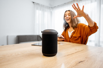 Wall Mural - Young cheerful woman controlling home devices with a voice commands, talking to a smart column at home. Concept of smart home and voice command control