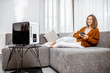 © rh2010 - Young woman relaxing on the couch while air humidifier or purifier working on the foreground. Controlling it with a smartphone remotely