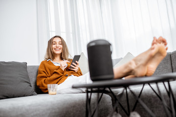 Wall Mural - Young cheerful woman controlling home devices with a voice commands, talking to a smart column while relax on the couch at home