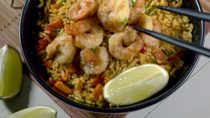 Wall Mural - Traditional shrimps stir fried with eggs, vegetables and rice in a black bowl on a bamboo placemat. Healthy Asian meal with sesame and lime. Someone’s hand takes a shrimp with chopsticks. 