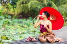 A Female Tourist Wearing A Thai Dress Is Taking Pictures With Lotus Flowers And Lotus Fields On A Wooden Bridge. Thai Woman Is Sitting On A Wooden Bridge Holding Red Umbrella And Pink Water Lilies.