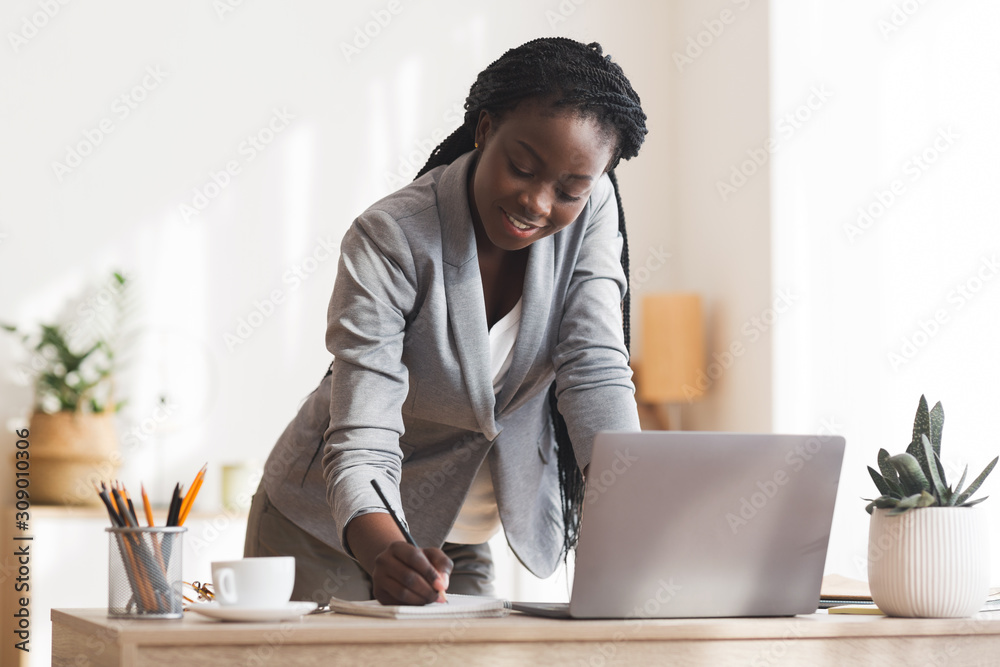 Busy black secretary taking notes while standing up near her desk - obrazy, fototapety, plakaty 