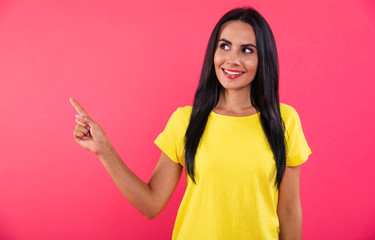 Sly smile. Close-up photo of a beautiful young woman in a yellow t-shirt, who is pointing to the left with her right index finger, looking to the left upper corner and smiling.