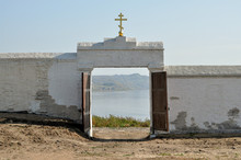 An Old White Brick Fortified Wall With Traces Of Repair And With An Open Gate In The Middle, A Golden Christian Cross Above Them, Through The Gate A View Of Land And Water And The Other Shore