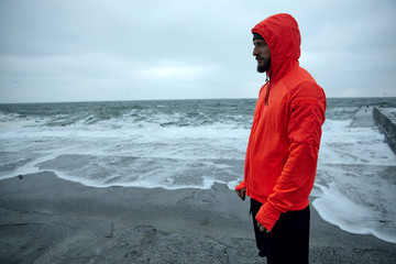 Wall Mural - Horizontal shot of young dark haired bearded sportsman dressed in warm orange hoodie and black athletic trousers standing over seaside on grey early morning and looking thoughtfully ahead