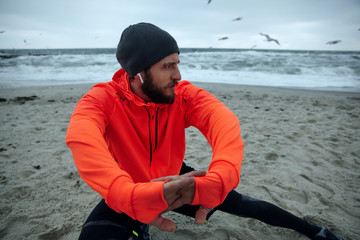 Wall Mural - Close-up of young serious brunette man with beard listening to music in his headphones while doing warm-up exercises before morning run at seaside, looking pensively aside and frowning face