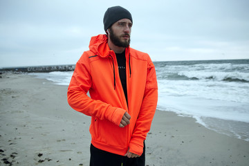 Wall Mural - Outdoor shot of young pensive bearded male looking thoughtfully ahaead while fastening zipper on his orange sporty hoodie, relaxing after morning workout