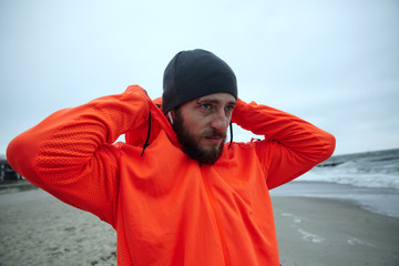 Wall Mural - Portrait of good looking young brunette man with lush beard and eyebrow piercing pulling on hood of his orange sporty coat while standing over seaside on gray early morning