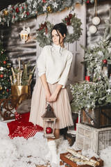 Wall Mural - Fashionable model girl in tulle skirt and knitted sweater holds a lantern in rustic interior decorated for New year. Young woman in modish winter outfit at exterior of house with Christmas decorations