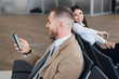 © Margo Kos - Young businessman and woman waiting at airport lounge. Man looking at phone. Business travelers waiting for their flight at airport terminal.