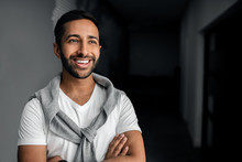Smiling Handsome Gen Z Indian Arabic Man Looking Into The Distance, Arms Crossed Wearing White T-shirt, Grey Cardigan Over His Shoulders