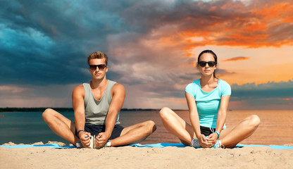 Canvas Print - fitness, sport and lifestyle concept - couple stretching legs on beach before training over sea and sunset sky on background