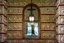Faro, Algarve / Portugal - Skulls And Bones Are Embedded In The Wall, A Lantern Is Lit, A Crucifix Behind A Glass Door, The Capella Chapel Of The Bones (Capella Dos Ossos), Church Of Carmo
