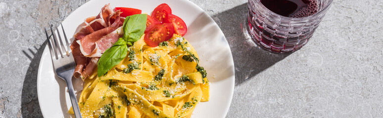 Wall Mural - top view of tasty Pappardelle with tomatoes, pesto and prosciutto with fork near red wine on grey surface, panoramic shot