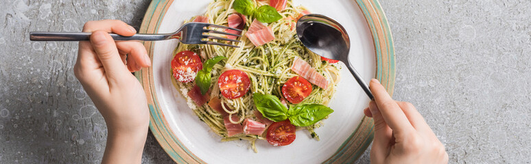 cropped view of woman eating Pappardelle with tomatoes, basil and prosciutto on grey surface, panoramic shot