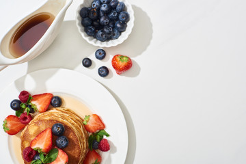 Wall Mural - top view of delicious pancakes with blueberries and strawberries on plate near maple syrup in gravy boat on marble white surface