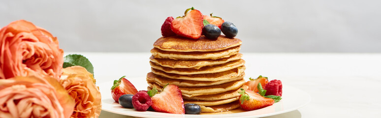 Wall Mural - selective focus of delicious pancakes with blueberries and strawberries on plate near rose flowers on white surface isolated on grey, panoramic shot