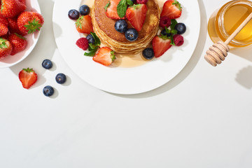 Wall Mural - top view of delicious pancakes with honey, blueberries and strawberries on plate on marble white surface