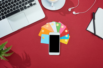 Wall Mural - top view of multicolored empty credit cards on red background with smartphone, laptop, earphones and coffee