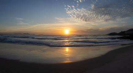 Wide panorama of waves coming in at the Arpoador Devil's beach in Rio de Janeiro, Brazil, at sunrise with the sun reflecting in the water that hits the pristine sand beach