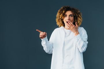 shocked stylish curly teenager in total white outfit pointing with hand isolated on green