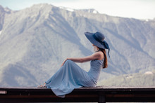Rear View Of A Slim Beautiful Young Woman In A Hat Sitting On A Bench Overlooking A Gorgeous Mountain Landscape And Admiring It On A Sunny Warm Summer Day While Relaxing. Travel Concept