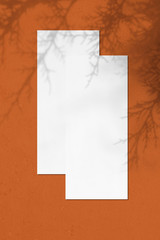 Poster - Two empty white vertical rectangle price-list or menu mockups with soft thuja leaves shadows lying on top of each other on orange concrete background. Flat lay, top view, copy space.