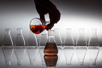 Hand pouring liquid in scientific laboratory glass erlenmeyer flask with glassware equipment on reflective surface.