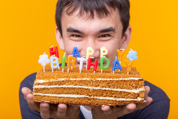 Wall Mural - Close-up of crazy cheerful young asian guy with glasses holding a burning candle in his hands and a congratulatory homemade cake on a yellow background. Birthday and anniversary celebration concept.