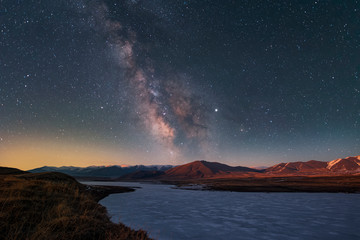Wall Mural - Beautiful starry night landscape. The bright galaxy of the Milky Way over a frozen lake and orange mountains lit by the light of sunrise.