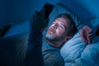 © TheVisualsYouNeed - happy man with blue eyes lying on bed late at night in dark light networking on mobile phone or online dating smiling relaxed enjoying internet in social media addiction